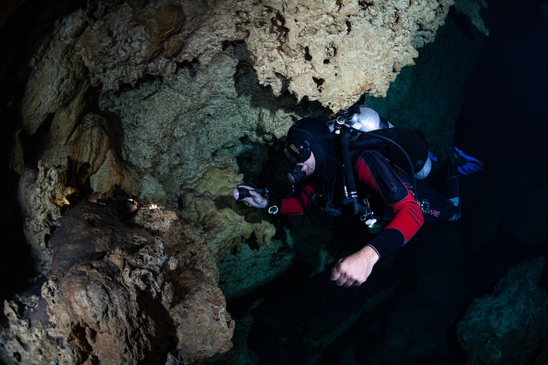 Cave diver in a red suit studies cenote stalactites, torch beam tracing the limestone.