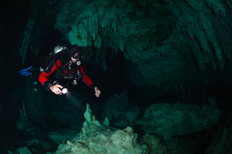 Torch beam cuts through a narrow cenote passage lined with stalactites and stalagmites.