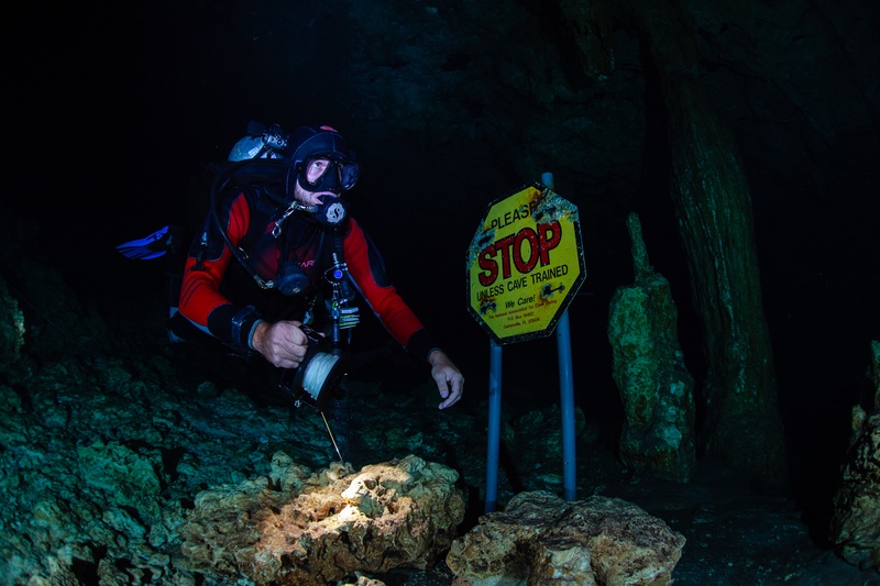 Red-suited cave diver kneels beside a yellow Stop Unless Cave Trained sign at the cenote restriction.