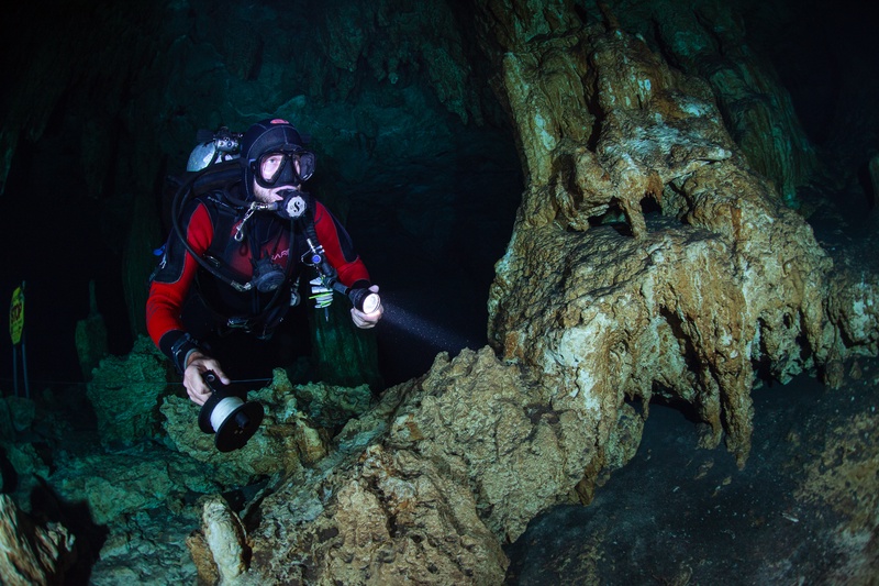Cave diver threads a reel through a cenote passage, torch beam skimming eroded limestone walls.