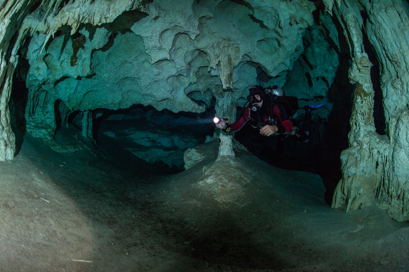 Cave diver threads a narrow stalactite passage, torch beam catching flowstone in the cenote.