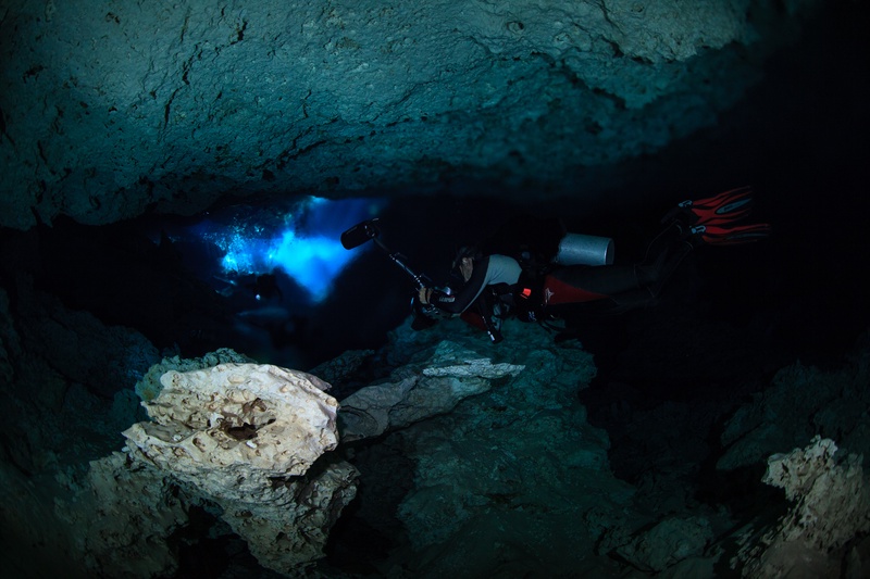 Cave diver slipping through a narrow cenote tunnel, torchlight catching the limestone walls.