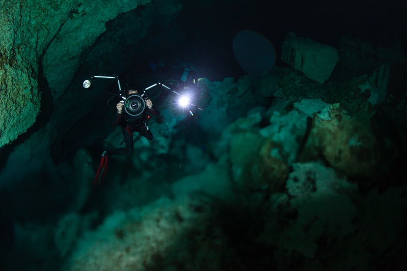 Cave photographer threads a narrow cenote passage, camera and strobes tucked between limestone walls.