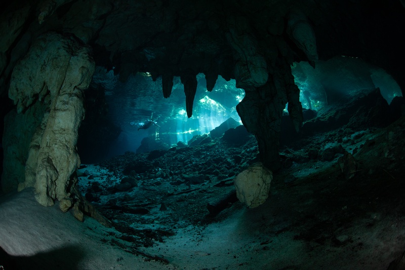 Stalactites and limestone columns frame a distant diver as light beams cut through the cenote cavern.