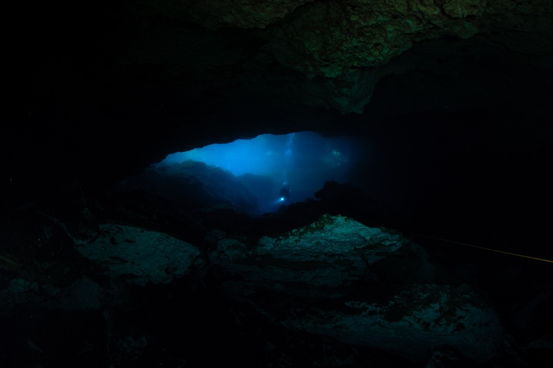 Blue cenote passage narrowing into darkness, a distant diver's torch glowing between limestone walls.