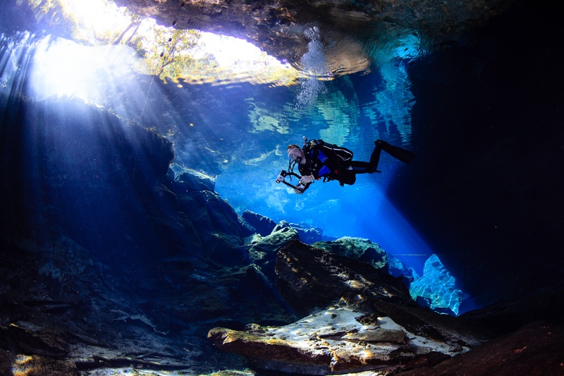 Underwater photographer hovers in the cenote as sunbeams pour through the opening above.