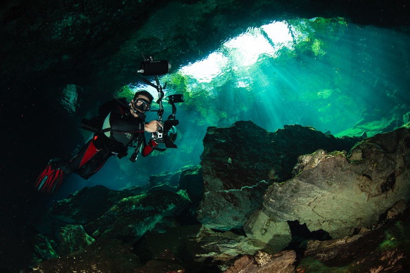 Photographer finning through a cenote cavern as sunbeams pour down from the opening above.