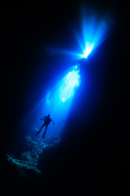 Diver silhouette hangs beneath a blue cenote light shaft, swallowed by the surrounding dark.