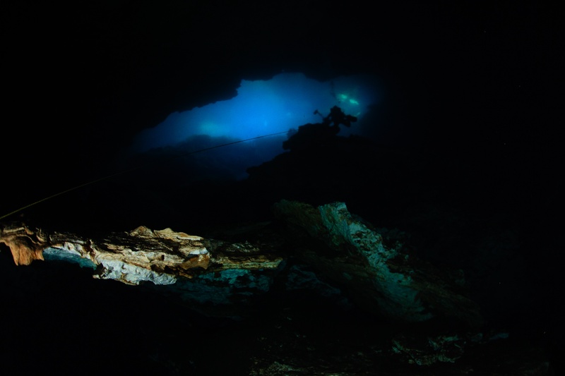 Cave diver moving toward a blue-lit cenote exit, dark rock formations framing the foreground.
