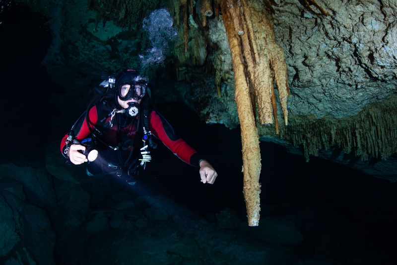 Diver hovers beside cenote stalactites, bubbles climbing toward the cavern ceiling overhead.