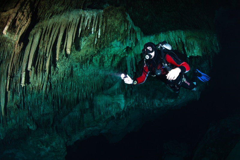 Cave diver in red and black finning past dense stalactites inside a flooded cenote.