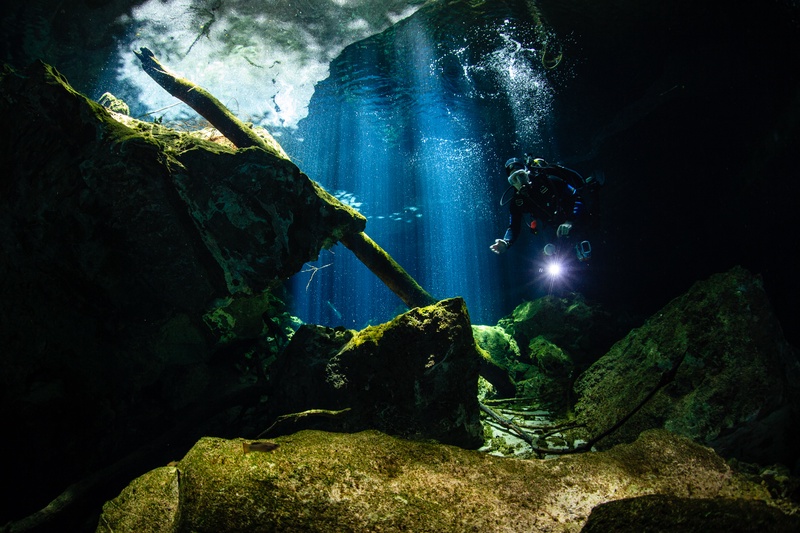 Torch beam leading the way, a cave diver threads between cenote boulders under blue shafts of light.