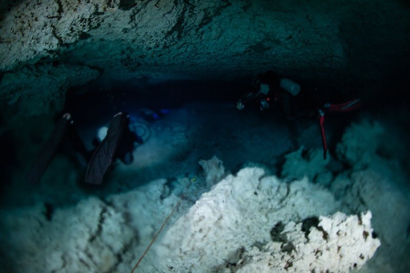 Two cave divers squeeze through a tight cenote restriction, torch beams scraping the limestone walls.