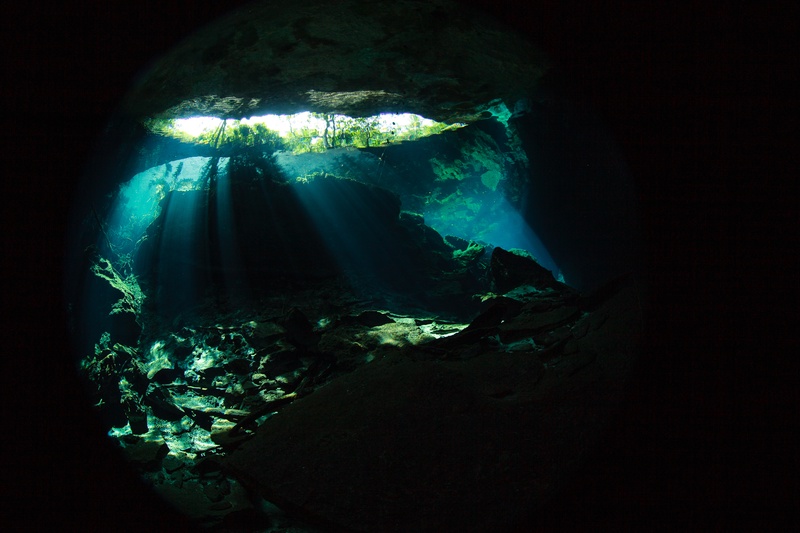 Light pours through a long cenote slit, catching roots, rock, and the cave floor below.