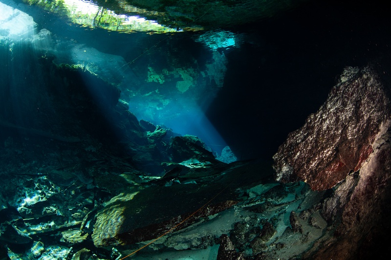 Sunbeams pour through the cenote opening, lighting boulders and sand on the cavern floor.