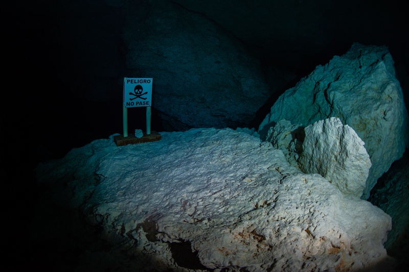 Peligro No Pase sign rests on pale limestone in a cenote cave, skull and crossbones stark underwater.