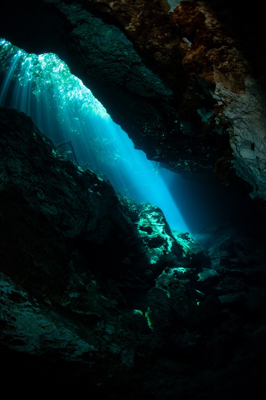 Turquoise light shaft dropping through a narrow cenote crack between rough limestone walls.