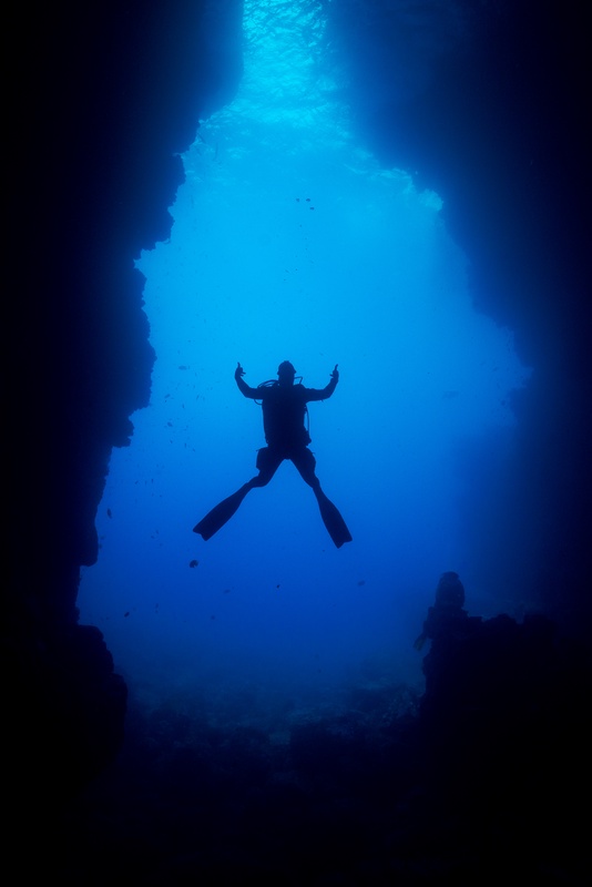 Scuba diver silhouetted arms-wide between towering underwater rock walls, blue light streaming from above.