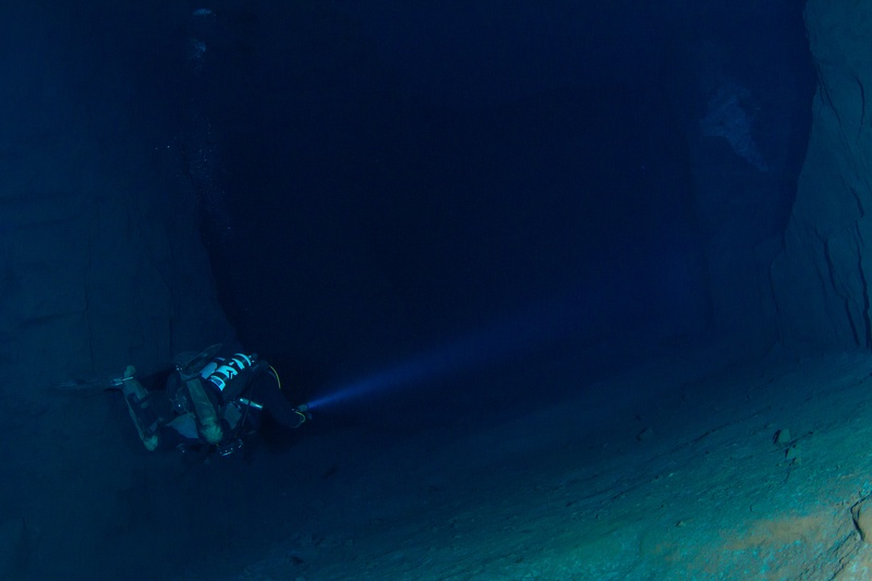 Technical diver descending along a vertical rock wall with torch beam cutting through the deep blue water of Kleinenbremen mine shaft.
