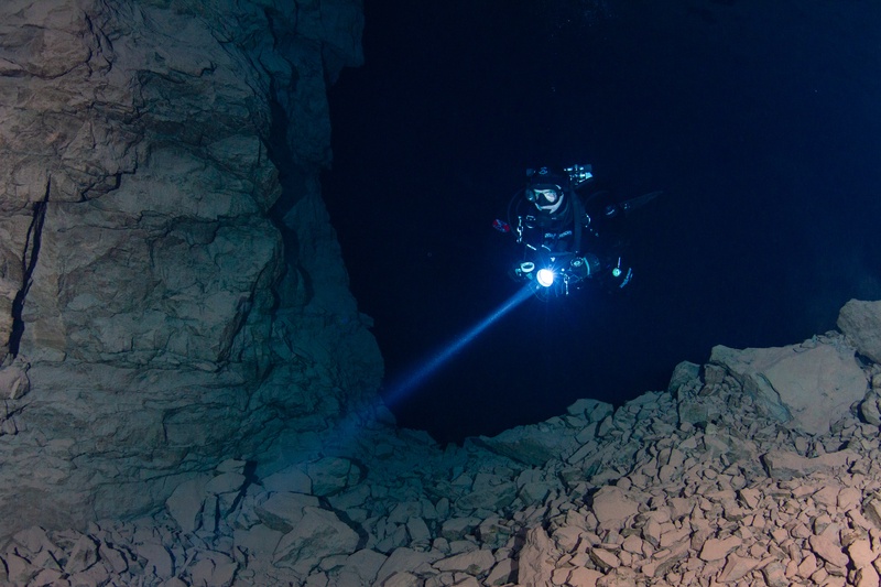 Cave diver with torch beam framed inside a narrow rocky portal opening into the flooded underground chambers of Kleinenbremen mine.